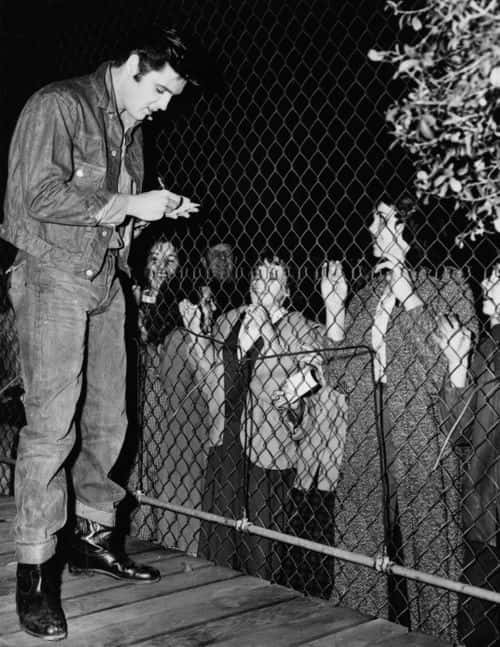 Elvis Presley (1935 - 1977) signing autographs for his fans, who are kept at bay by a wire fence.    (Photo by Keystone/Getty Images)