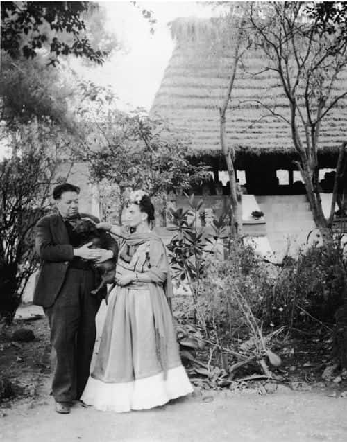Married Mexican painters Frida Kahlo (1907 - 1954) and Diego Rivera (1886 - 1957) stand together with a pet dog in front of thatchted-roof hut which houses a number of archeological artifacts, Mexico City, Mexico, 1940s. (Photo by Hulton Archive/Getty Images)