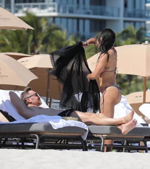 Jessica Ledon wraps up in black sheer overalls at the beach as partner David Guetta rests on the beach. (Backgrid)