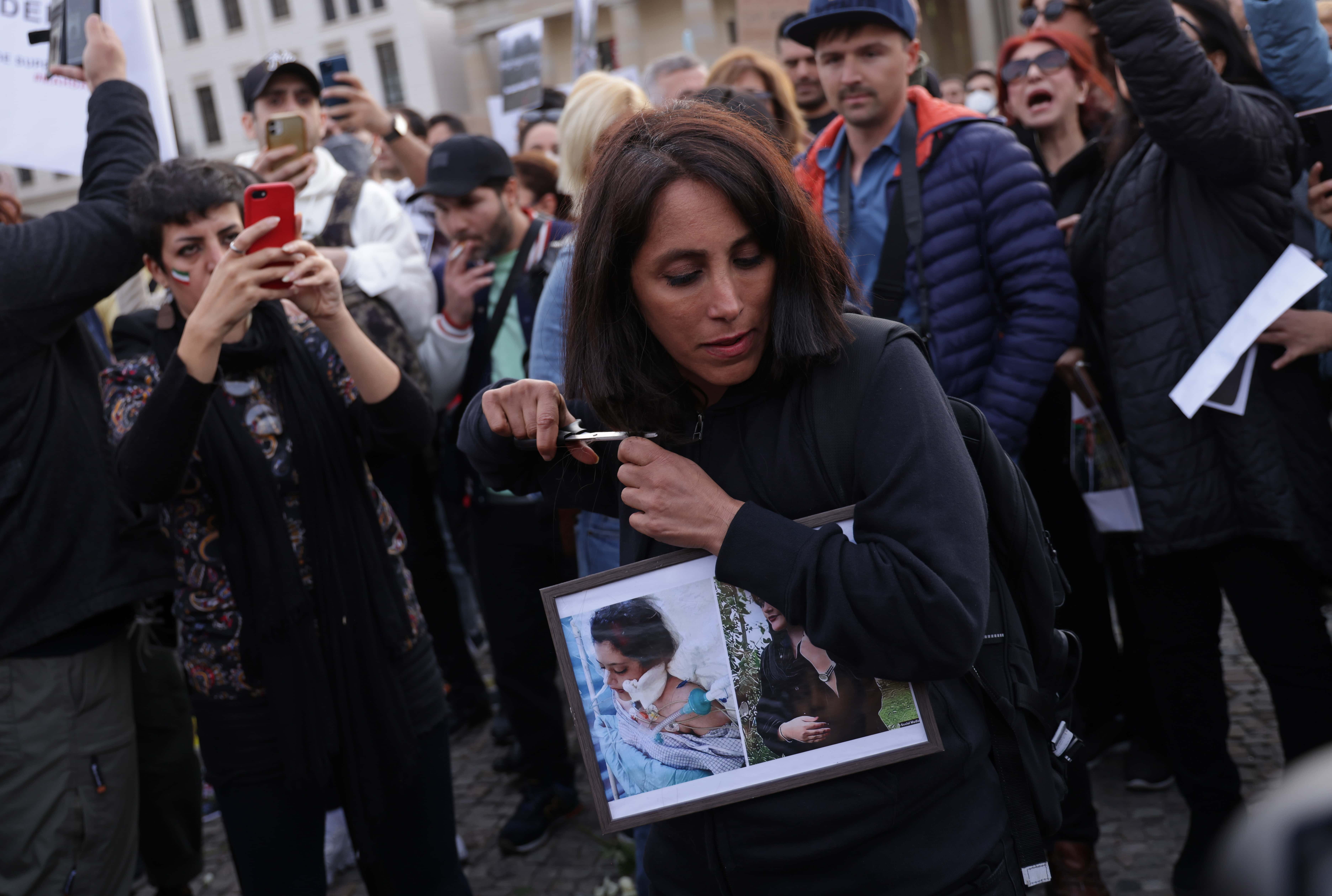 BERLIN, GERMANY - SEPTEMBER 23: A female protester cuts her hair with scissors as an act of solidarity with women in Iran while holding photographs of Mahsa Amini during a demonstration against the death Amini on September 23, 2022 in Berlin, Germany. Amini, 22, was arrested by Iranian authorities in Tehran on September 13 for not wearing her headscarf properly and died three days afterwards, apparently due to a severe head injury. Her death has sparked demonstrations across Iran nationwide that have spiralled into violence and left dozens of protesters dead. 
 (Photo by Sean Gallup/Getty Images)