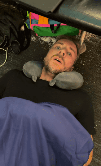 The camera then flips to Shepard, who is lying on the floor with a neck pillow and blanket (daxshepard/Instagram)