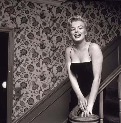 American actress Marilyn Monroe (1926 - 1962) stands in a staircase alongside a wall with a floral-motif pattern, late 1950s. She wears a black cocktail dress and dangling earrings. (Photo by Hulton Archive/Getty Images)