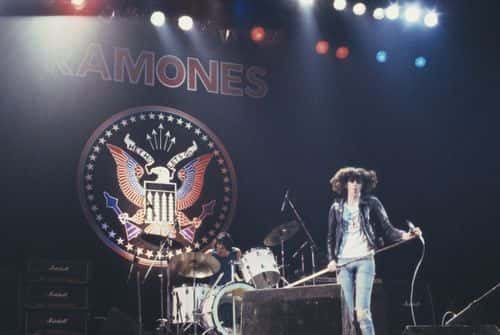 Joey Ramone (1951-2001), singer with US punk band the Ramones, on stage during a live concert performance by the band, with drummer Tommy Ramone in the background behind his drumkit, 1977. The backdrop has the band's 'presidential seal' logo and name. (Photo by Keystone/Hulton Archive/Getty Images)